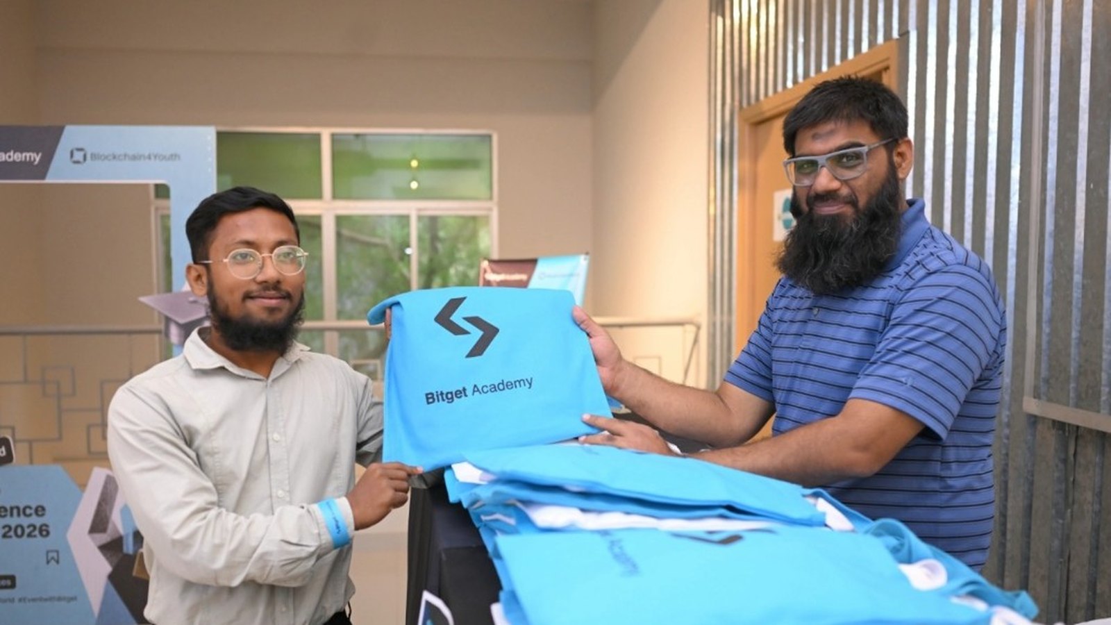 Event staff hold up a Bitget Academy T-shirt, with Blockchain4Youth materials visible in the background